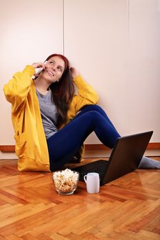 Young beautiful girl sitting on the floor in the bedroom in front of a laptop, preparing to watch a movie with popcorn and cup of coffee, talking on the phone.