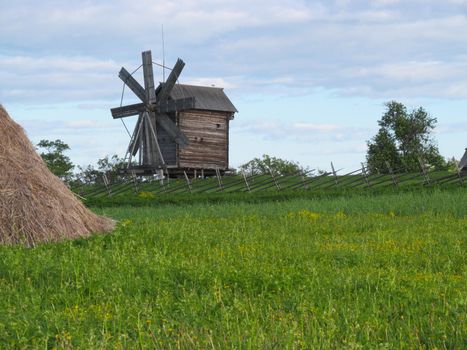 A wood mill against a blue sky with a green field in front of it on a bright Sunny day.