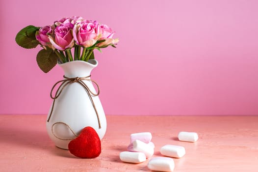 Small white and pink marshmallows are scattered on a pale pink background next to a vase of roses. Place for text.