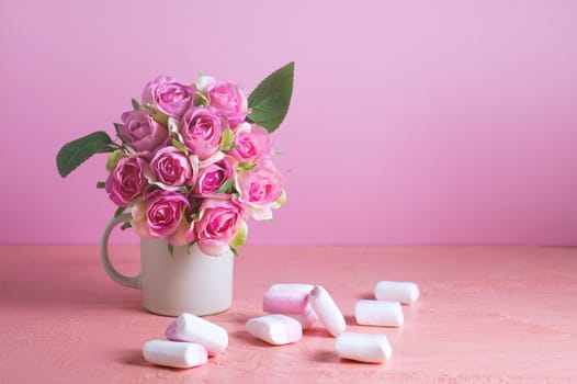 Small white and pink marshmallows are scattered on a pale pink background next to a vase of roses. Place for text.