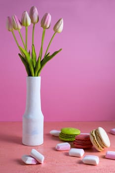 Macaroons and small white and pink marshmallows are scattered on a pale pink background next to a vase of tulips. Place for text.
