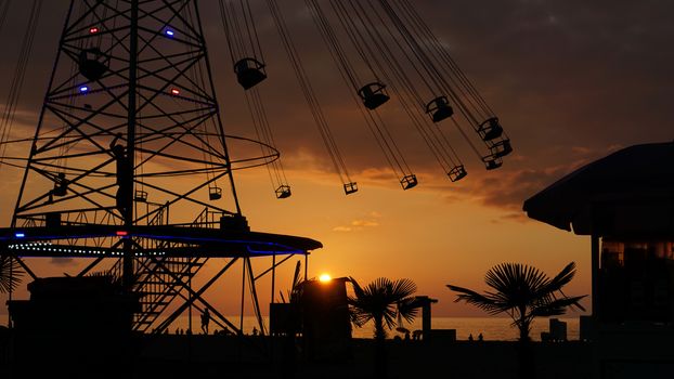 Swinging carousel roundabout chain ride at sunset. Entertainment on the beach, silhouettes of palm trees on a background of sea sunset