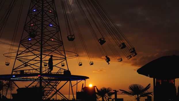 Swinging carousel roundabout chain ride at sunset. Entertainment on the beach, silhouettes of palm trees on a background of sea sunset