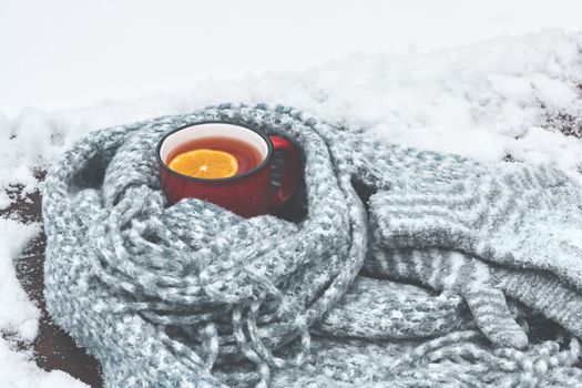 Red enameled cup of hot tea with lemon wrapped in a knitted scarf on a snowy wooden table.