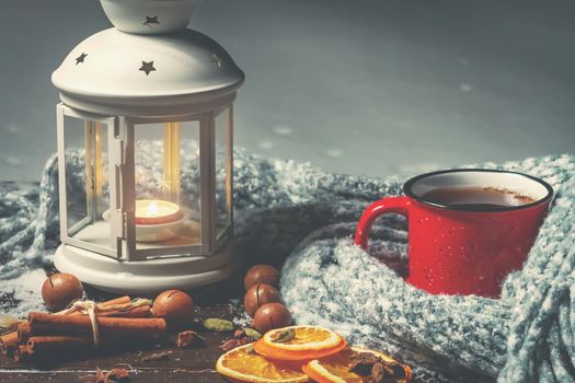 Lantern with a burning candle, spices and a red mug with hot coffee on a snowy wooden table.