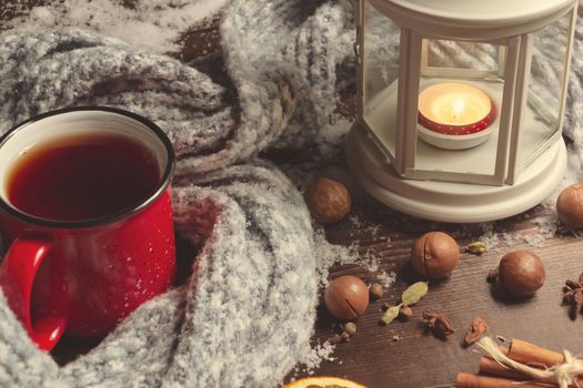 Lantern with a burning candle, spices and a red mug with hot coffee on a snowy wooden table.