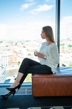 Young businesswoman drinking coffee in office near panoramic window