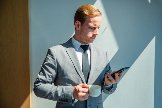 Typing business message. Confident young man in suit drinking coffee and holding mobile phone in office