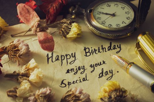 Happy birthday enjoy your special day. Vintage brass pen writing birthday greetings on old paper. Decoration by pocket watch and dry flower around it.