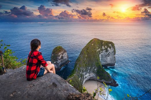 Woman sitting on cliff and looking at sunset at Kelingking Beach in Nusa penida island, Bali, Indonesia.