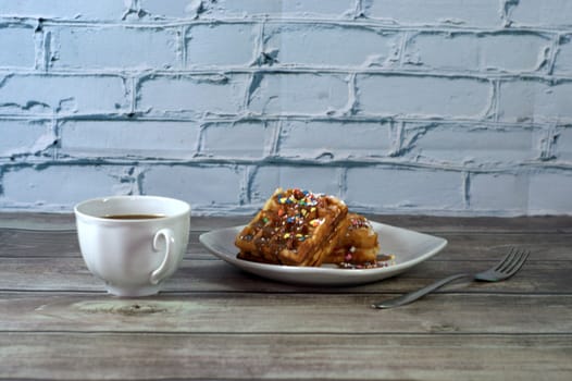 Breakfast, a fork, a cup of black coffee and a plate with two Viennese waffles are on a wooden table. Close-up.