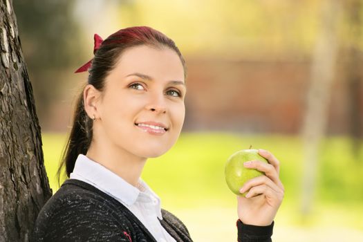 Healthy eating outdoors. Beautiful smiling young girl holding an apple, in the park leaning against a tree.