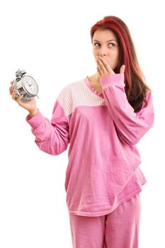 Oops, I overslept again. Amused beautiful young girl in pink pajamas holding an alarm clock, secretly giggling, isolated on white background.