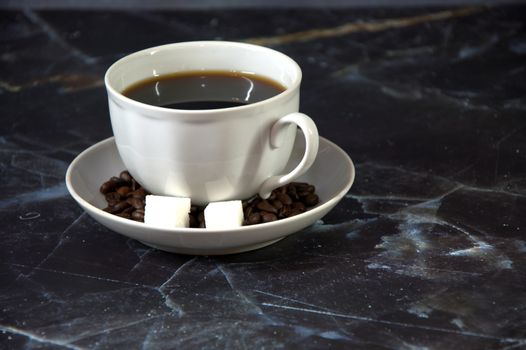 A white ceramic cup of black coffee on a saucer with two cubes of sugar and sprinkled coffee beans. Close-up.