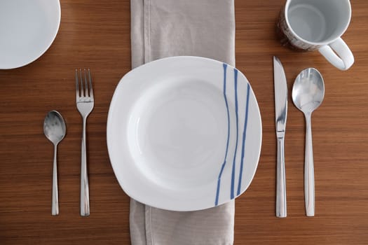 White porcelain tableware with bright metal cutlery and soft napkin on a wooden table.