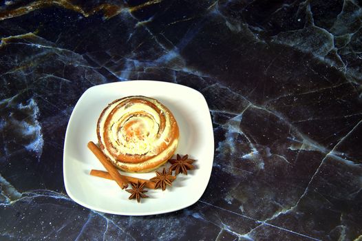 White ceramic plate with a bun with cinnamon in the glaze, stars anise star and cinnamon sticks on a marble background. Close-up.