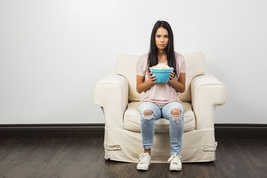 young millenial girl, holding a blue bowl of popcorn, sitting on a white couch