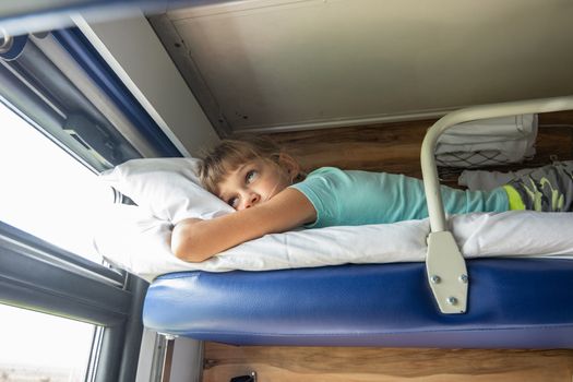 An eight-year-old girl lies on the top shelf of a reserved seat car and looks tiredly out the window