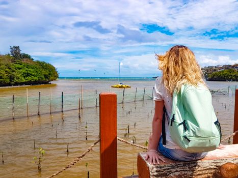 A girl with a backpack sits on a bench on the shore of Boracay island of the Philippines and looks at the thickets of mangroves in the water. Rear view. Follow me.