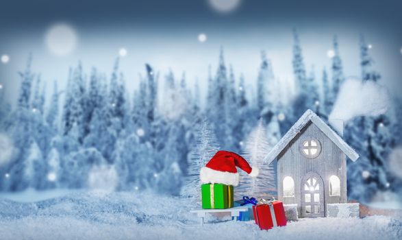 Christmas card with cozy small decorative house , gifts and Santa Claus cap on sleigh on snow over fairy tale winter fir forest background and falling snow . New Year design postcard . Copy space for text