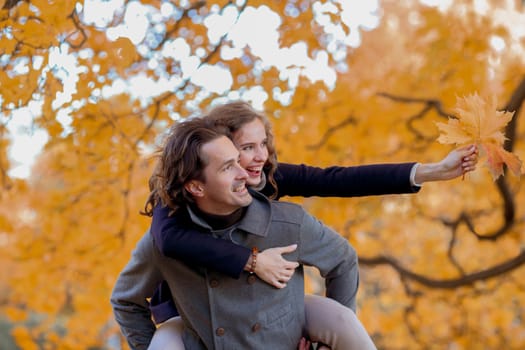 Couple in autumn park having fun, smiling man carrying woman piggyback outdoor