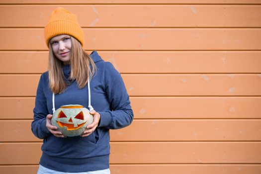 Woman in casual clothes and hat holding Halloween pumpkin over orange wall background with copy space for text