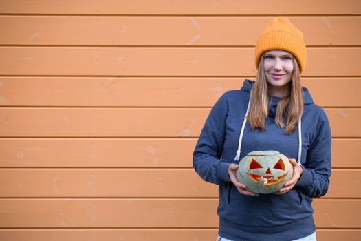 Woman in casual clothes and hat holding Halloween pumpkin over orange wall background with copy space for text