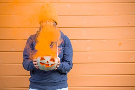 Woman in casual clothes and hat holding Halloween pumpkin over orange wall background with copy space for text