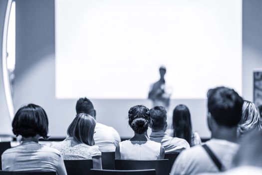 Speaker giving a talk in conference hall at business event. Focus on unrecognizable people in audience. Business and Entrepreneurship concept. Blue toned greyscale image.