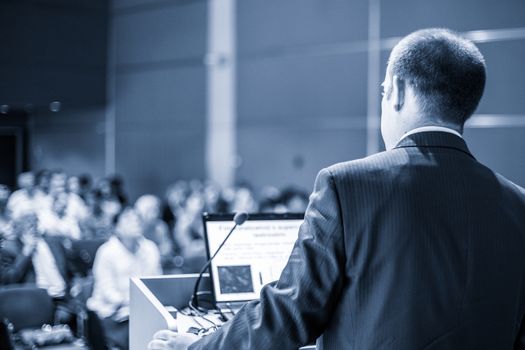 Speaker giving a talk on corporate business conference. Unrecognizable people in audience at conference hall. Business and Entrepreneurship event. Blue toned grayscale image.