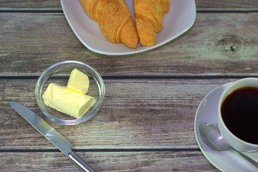 A traditional breakfast, a cup of black coffee, butter and a plate with two fresh croissants.