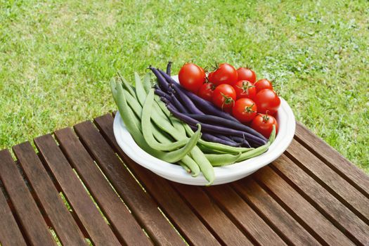 Freshly harvested Calypso beans, French beans and cherry tomatoes in a dish on a wooden picnic table in a garden