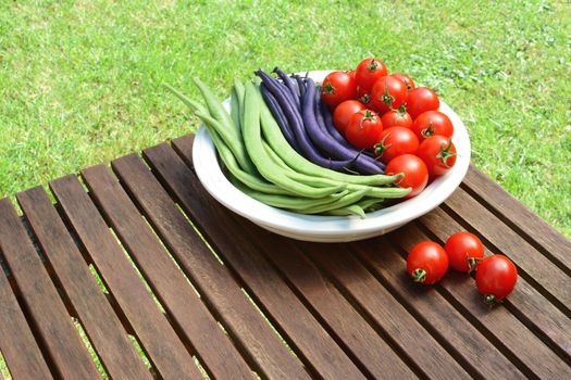 Green calypso beans and purple French beans in a dish with red cherry tomatoes, three spilled on the wooden picnic table 