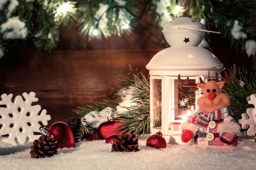 White lantern with a burning candle stands in the snow surrounded by Christmas decorations on the background of a wooden wall, Christmas tree branches and lights.