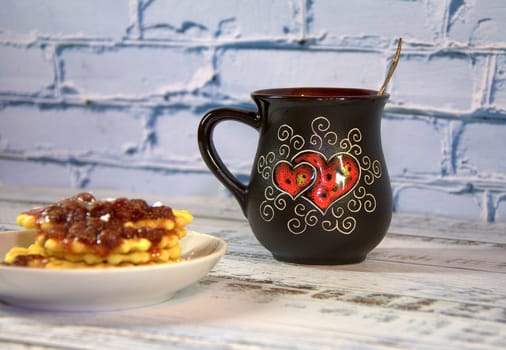 A dark ceramic mug with tea and a stack of waffles on a saucer with strawberry jam. Close-up.