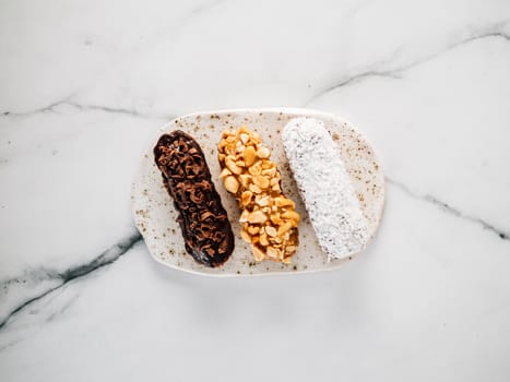 Set of three homemade eclairs on marble background. Top view of delicious healthy profitroles with different decor elements - chocolate, peanut and sherdded coconut
