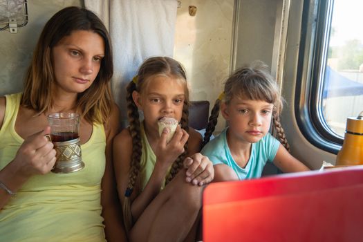 Mom and two girls are looking at the laptop screen in the train car, having a bite while