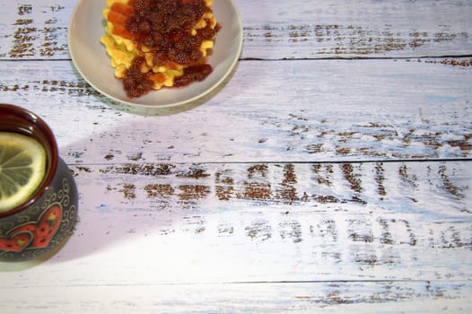 A mug of herbal tea with lemon and saucer with a stack of waffles with strawberry jam. Close-up.