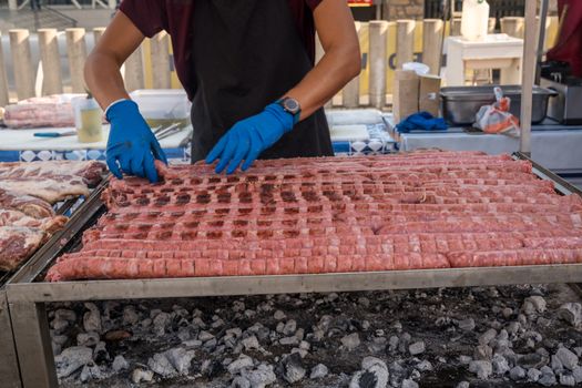 BBQ Sausage lined up on a large outdoor grill with smoke coming up from the fire below. person in black apron blue shirt gloved hand holding tongs turning meat