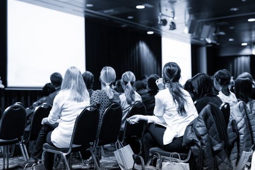 Business and entrepreneurship symposium. Speaker giving a talk at business meeting. Audience in conference hall. Rear view of unrecognized participant in audience.