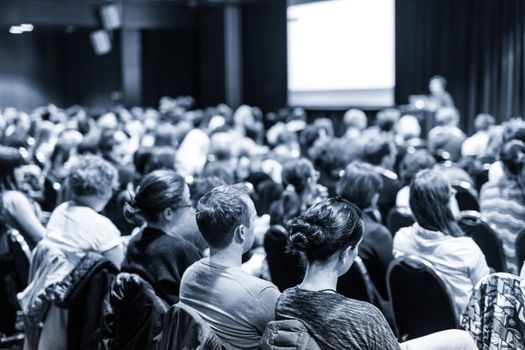 Business and entrepreneurship symposium. Speaker giving a talk at business meeting. Audience in conference hall. Rear view of unrecognized participant in audience.