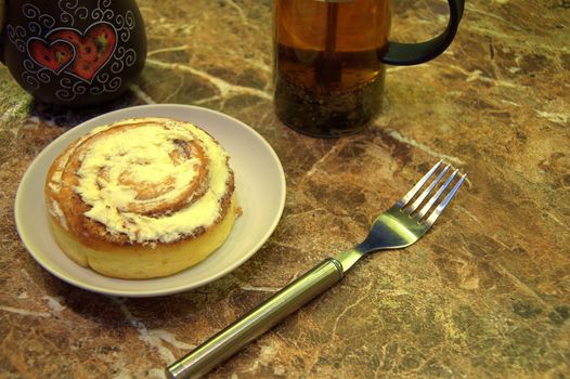 Cinnamon roll in icing on a saucer, a cup of tea, teapot and fork. Close-up.