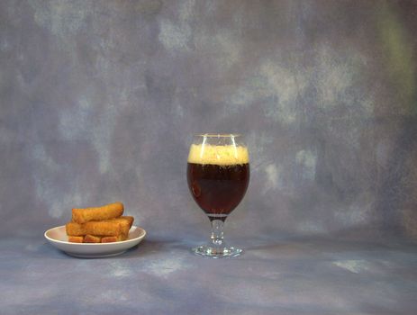 Glass of dark beer with foam and a plate with wheat croutons on a gray background. Close-up.