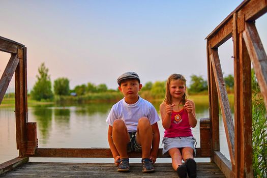 Cute children, little boy and girl are sitting on pier in the summer evening. Two siblings. Golden hour by the lakeside. Happy childhood and family concept. 