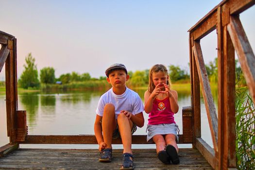 Children sitting on pier. Two children of different age - elementary age boy and preschool girl sitting on a wooden pier. Girl making heart shape. Summer and childhood concept. Children on bench at the lake.