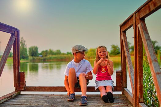 Children sitting on pier. Two children of different age - elementary age boy and preschool girl sitting on a wooden pier. Girl making heart shape. Summer and childhood concept. Children on bench at the lake.Golden hour and summer sunset.