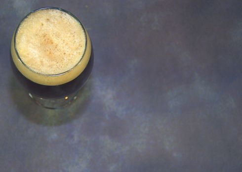 A glass of dark beer with foam on a gray background from a top angle. Close-up.