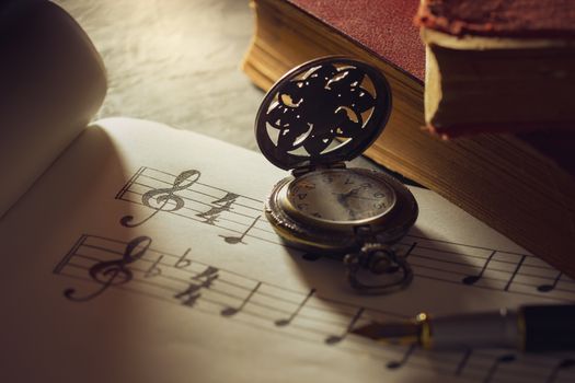 Music notes and old book with pocket watch on wooden table background in morning light. Writing chords by vintage pen. Closeup and copy space for text. Concept of Music lovers.