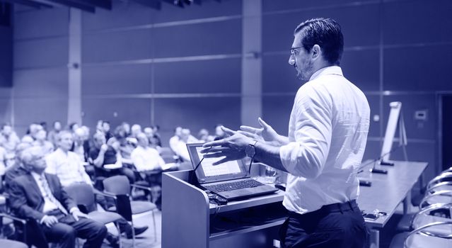 Speaker giving a talk on corporate Business Conference. Audience at the conference hall. Business and Entrepreneurship event. Greyscale blue toned image.