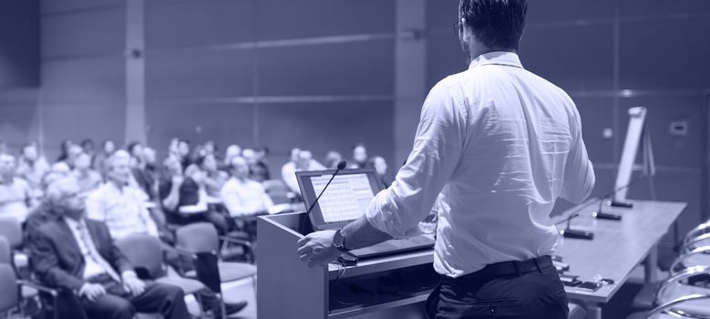 Speaker giving a talk on corporate Business Conference. Audience at the conference hall. Business and Entrepreneurship event. Panoramic composition. Greyscale blue toned image.